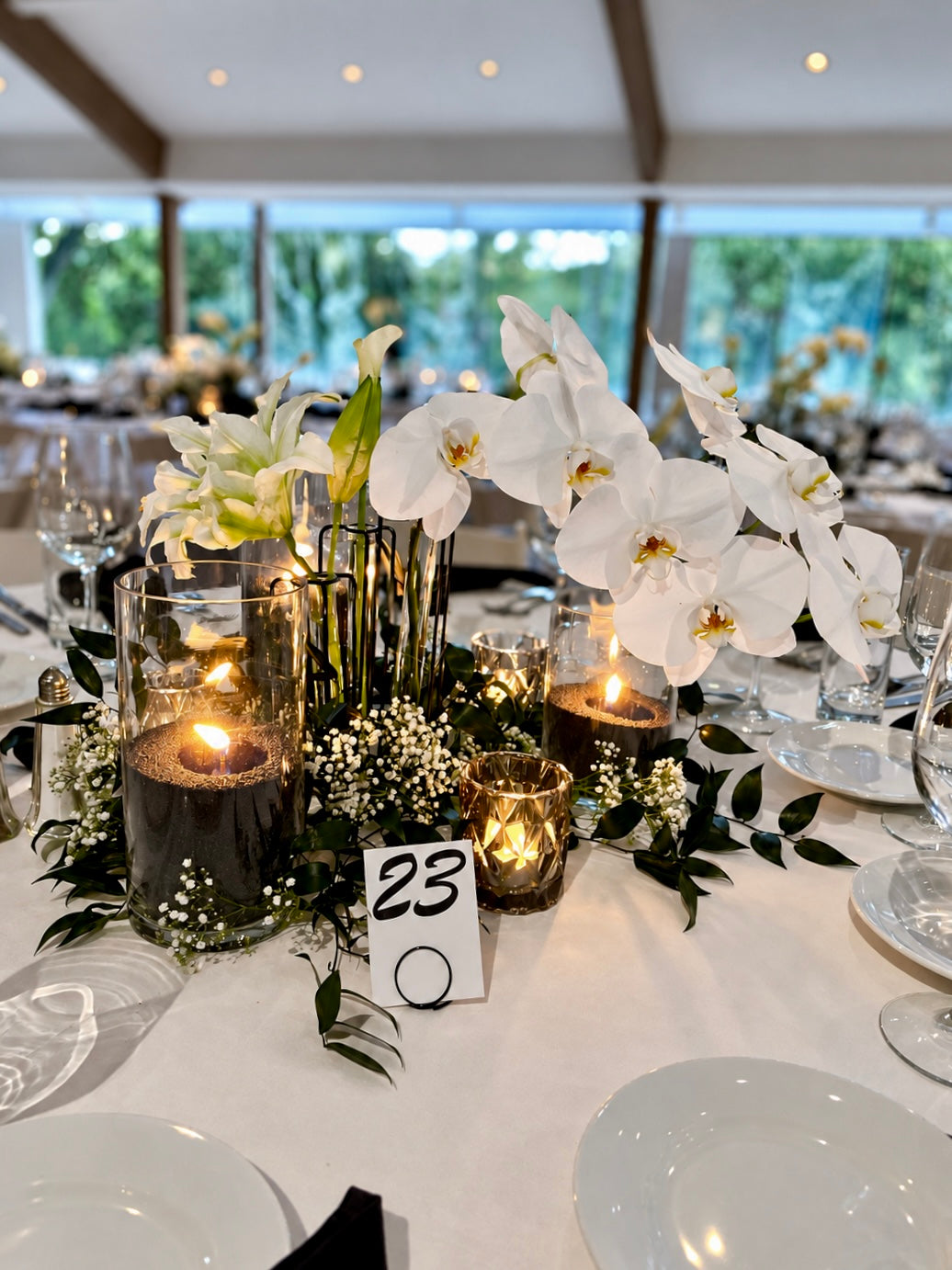 A styled wedding table modern centerpiece with white orchids, black foliage, and multiple pearled wax candles in black on a white tablecloth.