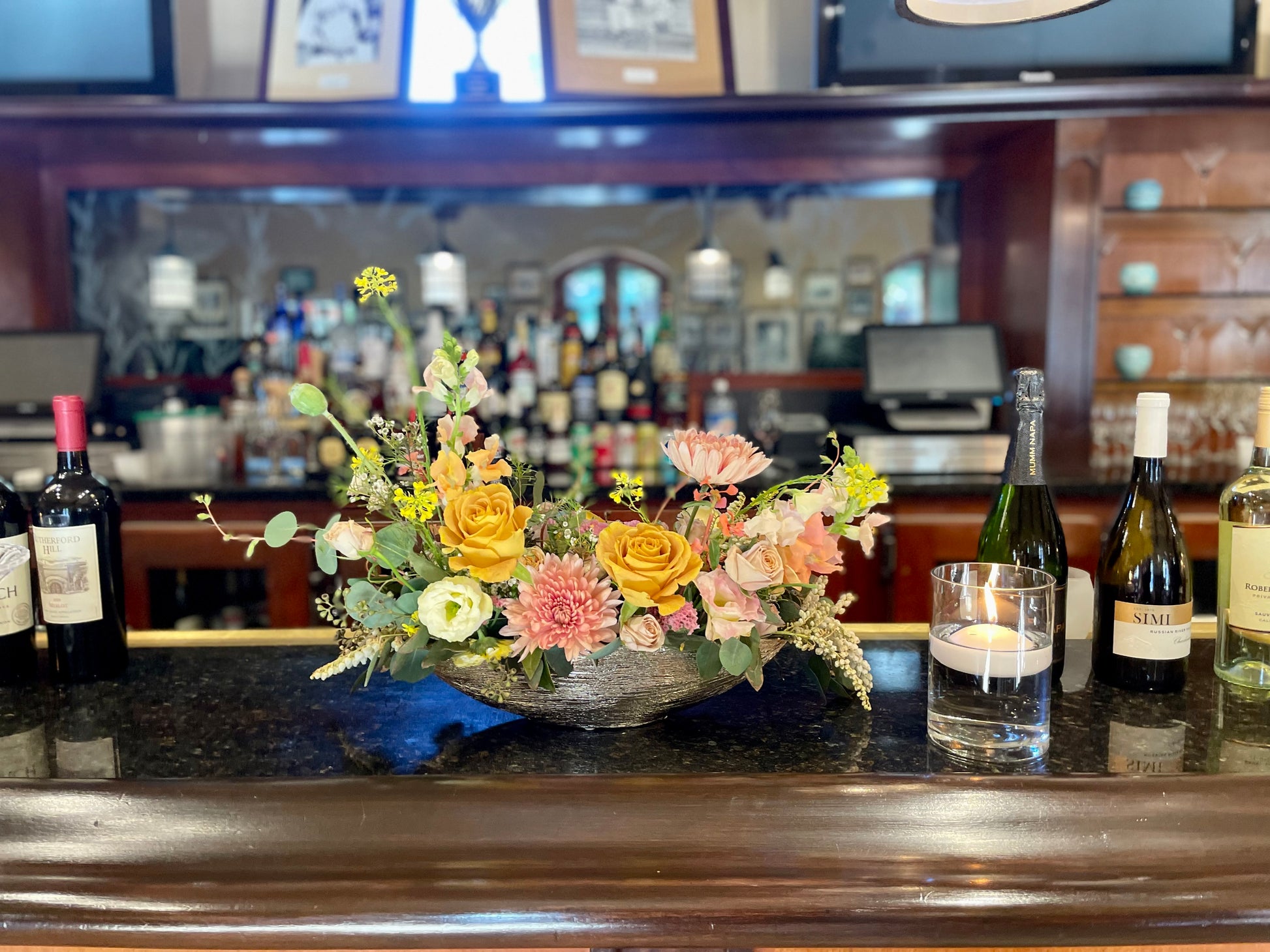 A vibrant floral centerpiece with yellow, pink, and orange flowers arranged in a dark bowl on a polished dark wood bar counter, with wine bottles and a glass candle holder in the background.