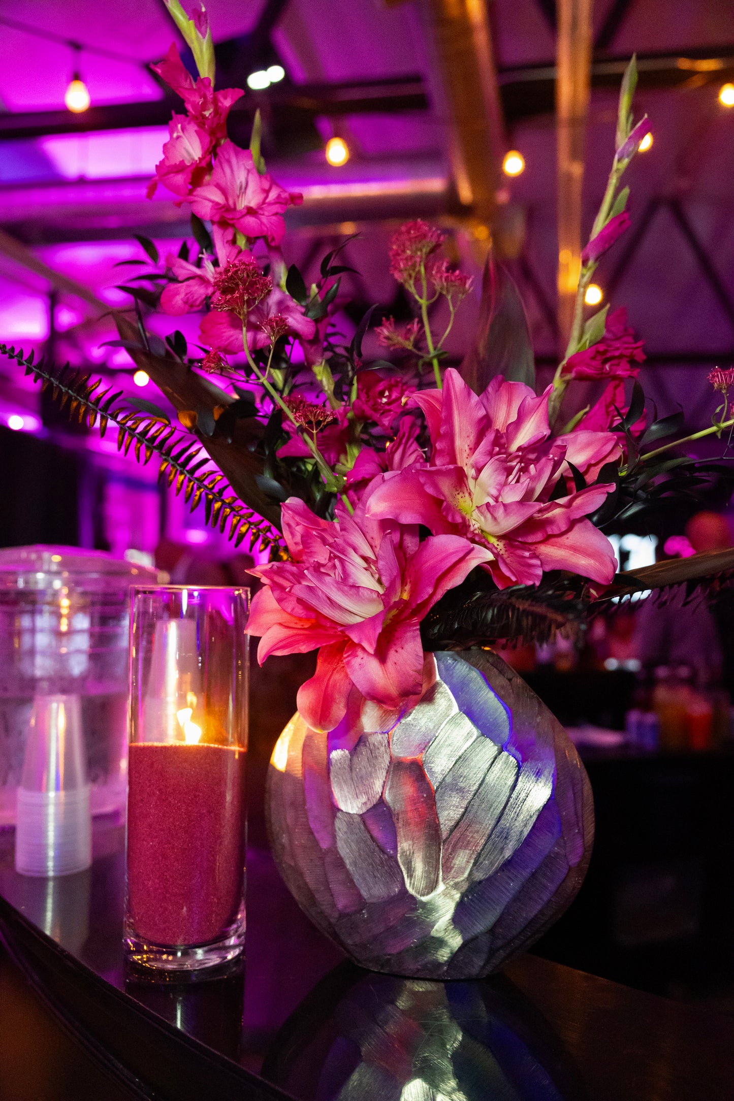 A bold, but minimalist floral centerpiece with pink and magenta flowers arranged in a textured metallic silver vase on a dark table, with a lit pink candle in a clear glass holder beside it. The background shows a dimly lit event space with purple ambient lighting and warm hanging lights.