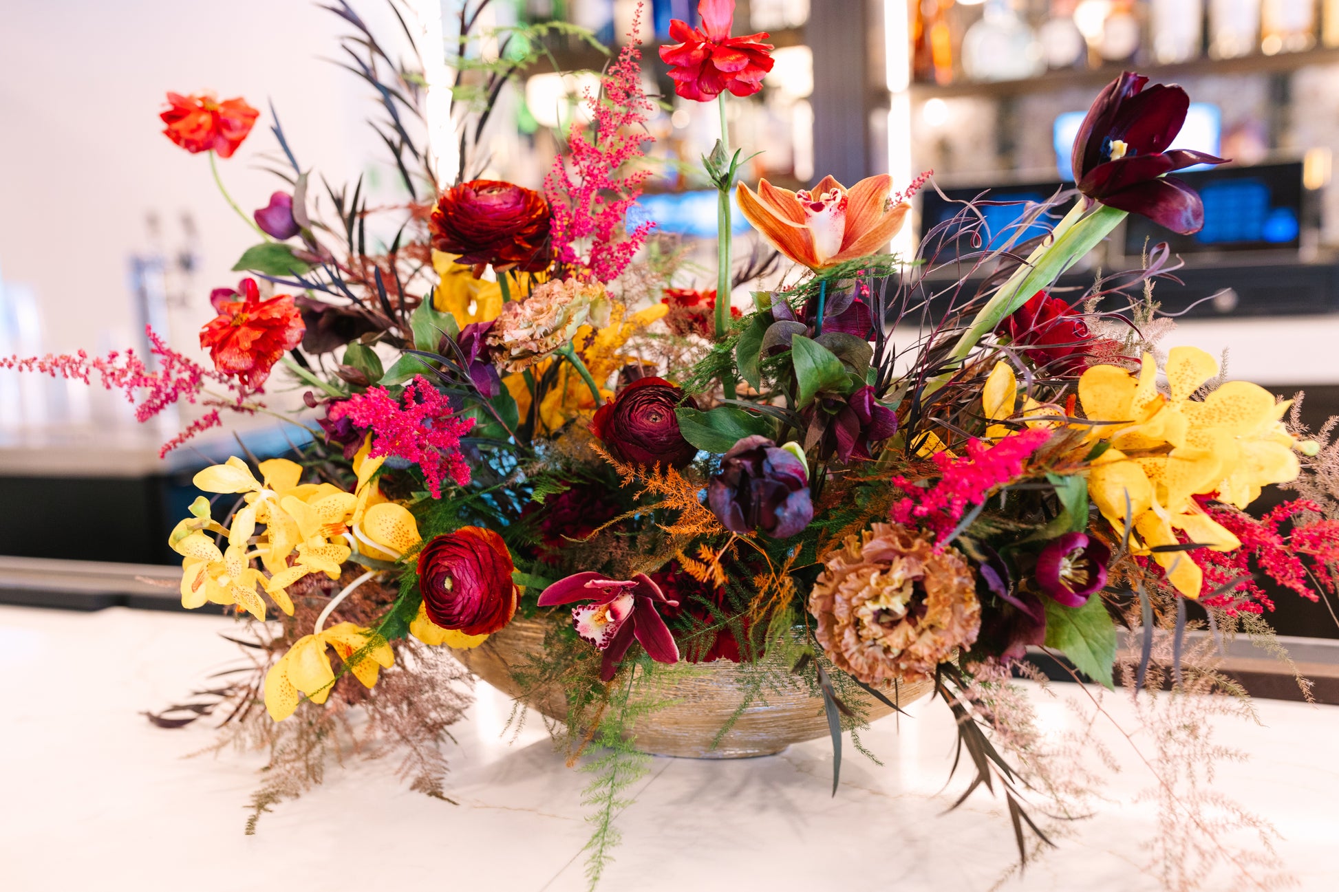 A vibrant, multicolored floral arrangement in a woven basket bowl, featuring red, yellow, orange, pink, and purple flowers with green foliage, set on a light-colored table with a blurred indoor background.