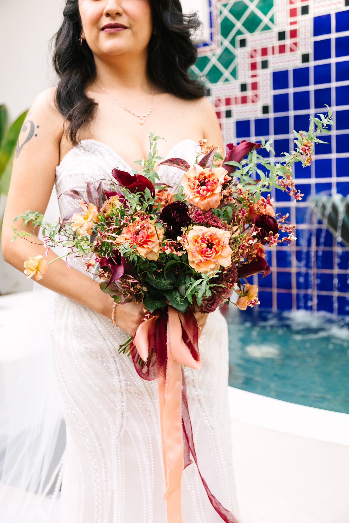 A woman in a white strapless gown holds a large floral bouquet with peach Charlotte Ranunculus, burgundy ranunculus and calla lilies, exotic orchids, spirea and butterfly ranunculus at a wedding at Spanish Hills Club in Camarillo, CA 93010.