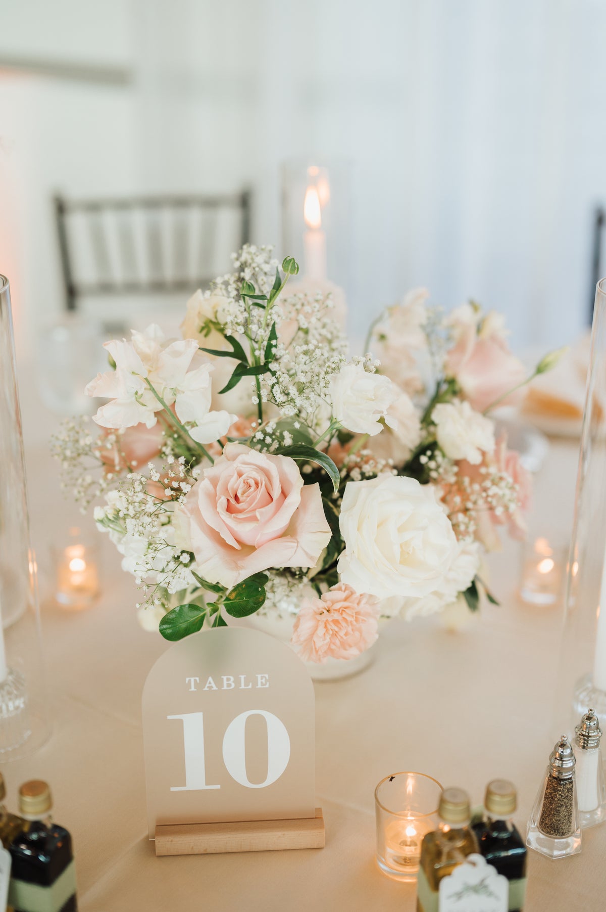 A floral centerpiece with blush pink and white roses, baby's breath, and green leaves on a table, with a clear acrylic table number sign reading 10 in front. Small candles and bottles are visible around the table in a softly lit indoor setting.