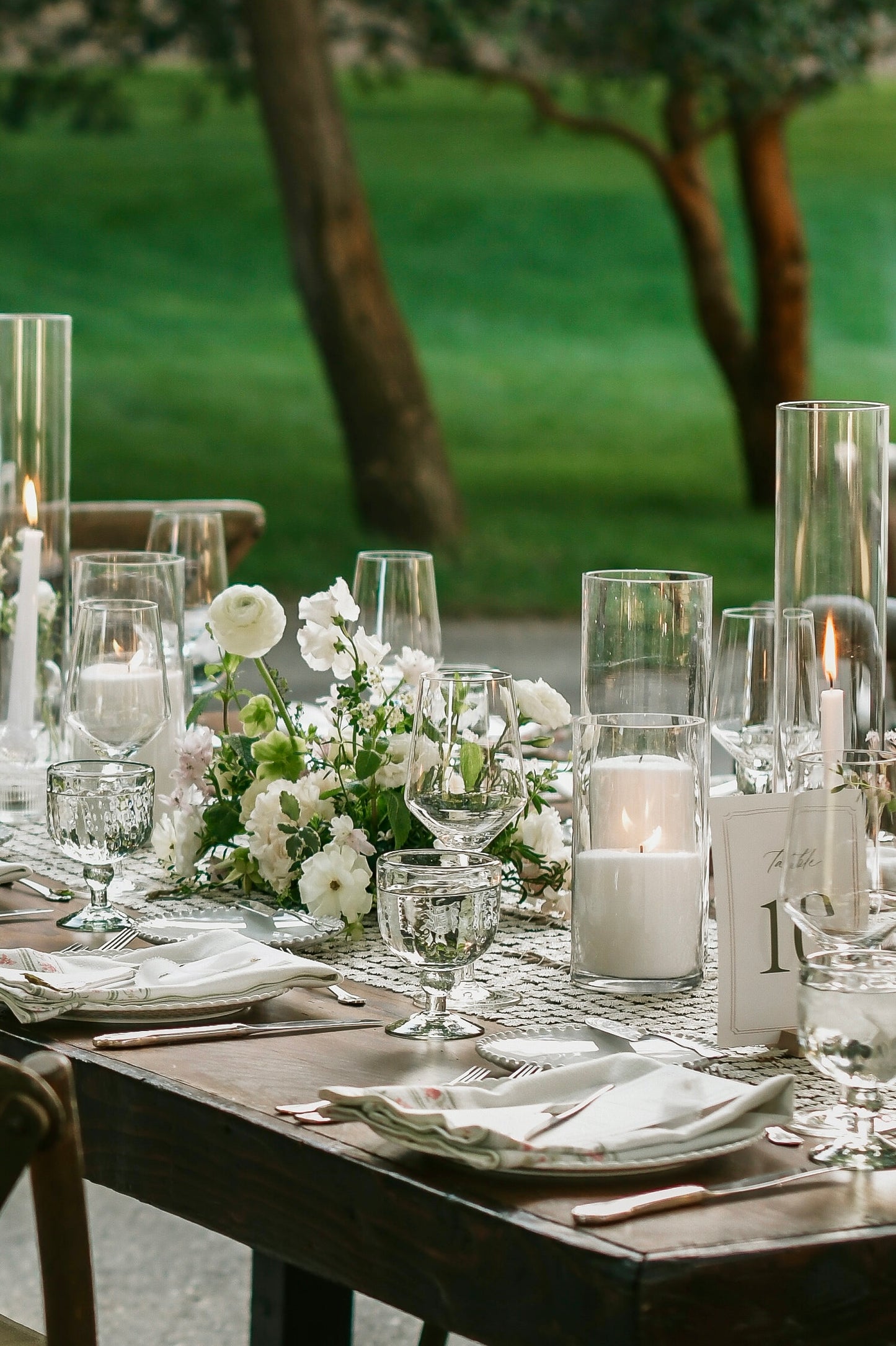 An outdoor wedding-style table setting with white flowers, clear glass candleholders, wine glasses, plates, folded white napkins, and lit white pillar candles, with a blurred green lawn and trees in the background.