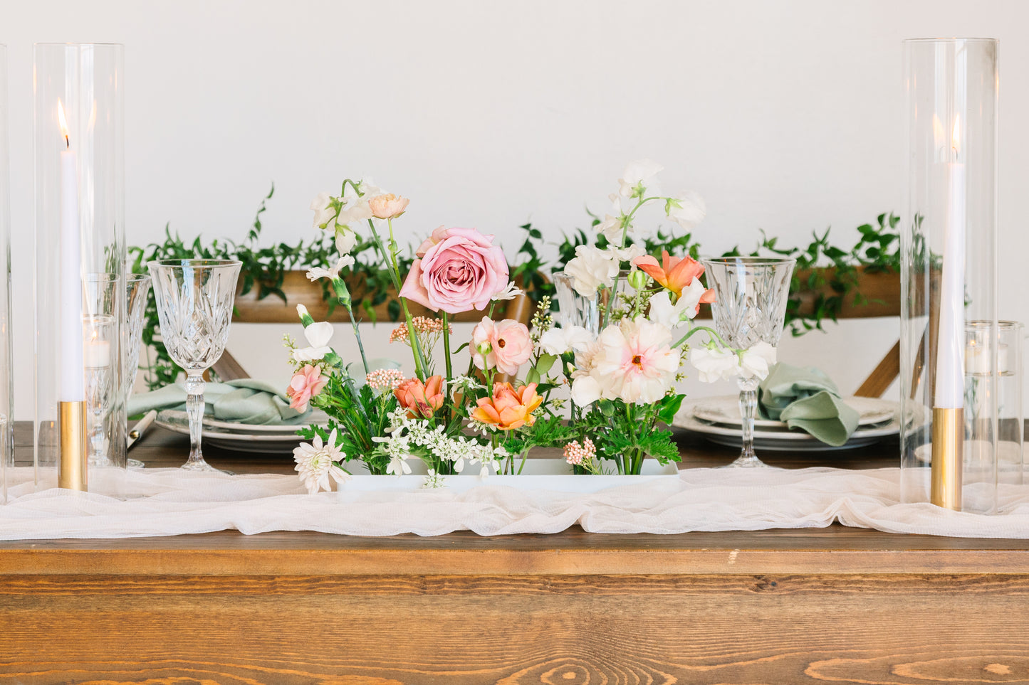 An ikebana style, modern, minimalist centerpiece with pink, peach, and white flowers on a blush table runner, flanked by elegant taper candles set on a wooden farm table.