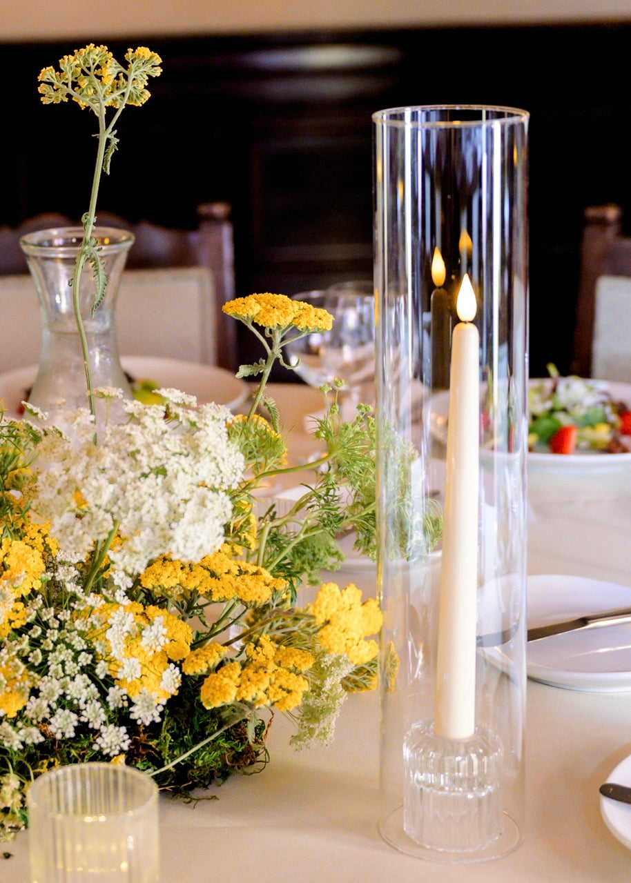 A tall, clear cylindrical glass candle holder with a realistic LED taper candle, placed on a dining table beside a floral centerpiece with yellow and white flowers at a wedding at the Ebell of Los Angeles.