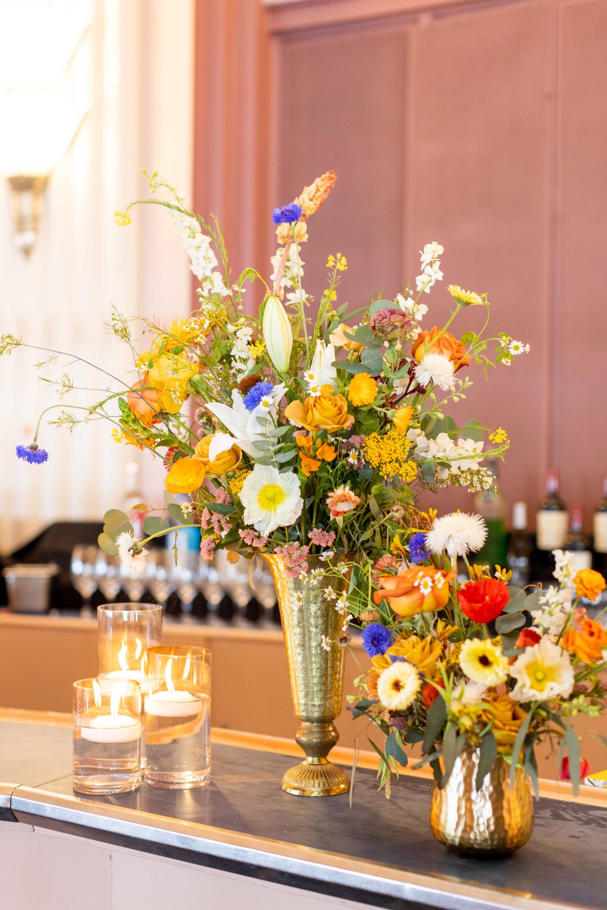 A large, vibrant floral centerpiece arrangement in a tall metallic gold vase on a bar counter, alongside two lit tealight candles and a smaller matching floral arrangement in a gold pot.