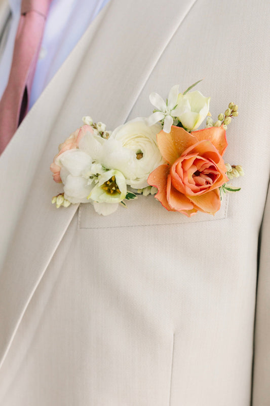 Floral pocket square boutonniere featuring a white ranunculus and coral spray rose on a light-colored suit lapel