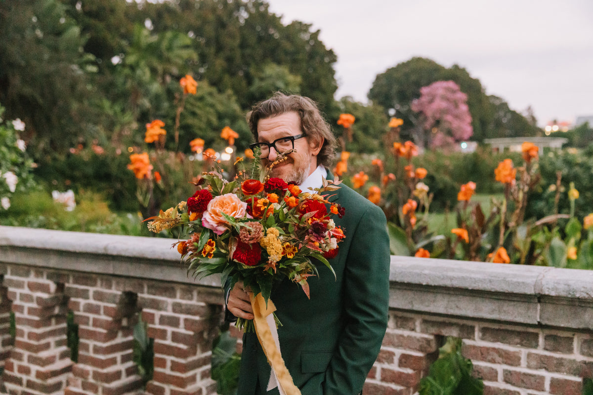 A groom wearing a dark green suit holding a large, vibrant fall wedding bouquet of red, orange, and yellow flowers at The Natural History Museum at their rose garden ceremony setting.