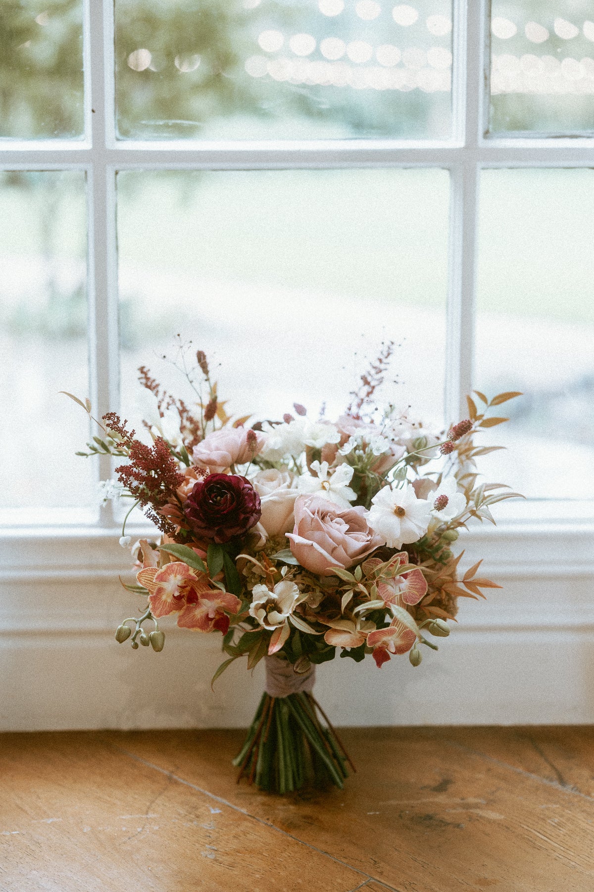 A textural, wedding bouquet in burgundy, dusty rose, cream and soft rust tones, resting on a wooden floor in front of a bright window at The Lodge at Malibou Lake in Agoura Hills. 