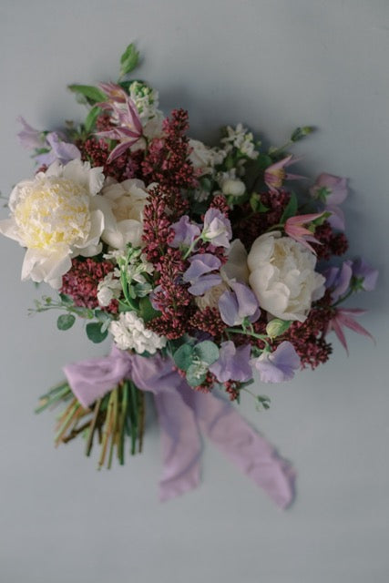 A garden-style floral bouquet featuring white peonies, light purple sweet peas, fresh and fragrant lilac, and delicate eucalyptus foliage, tied with a lavender silk ribbon.