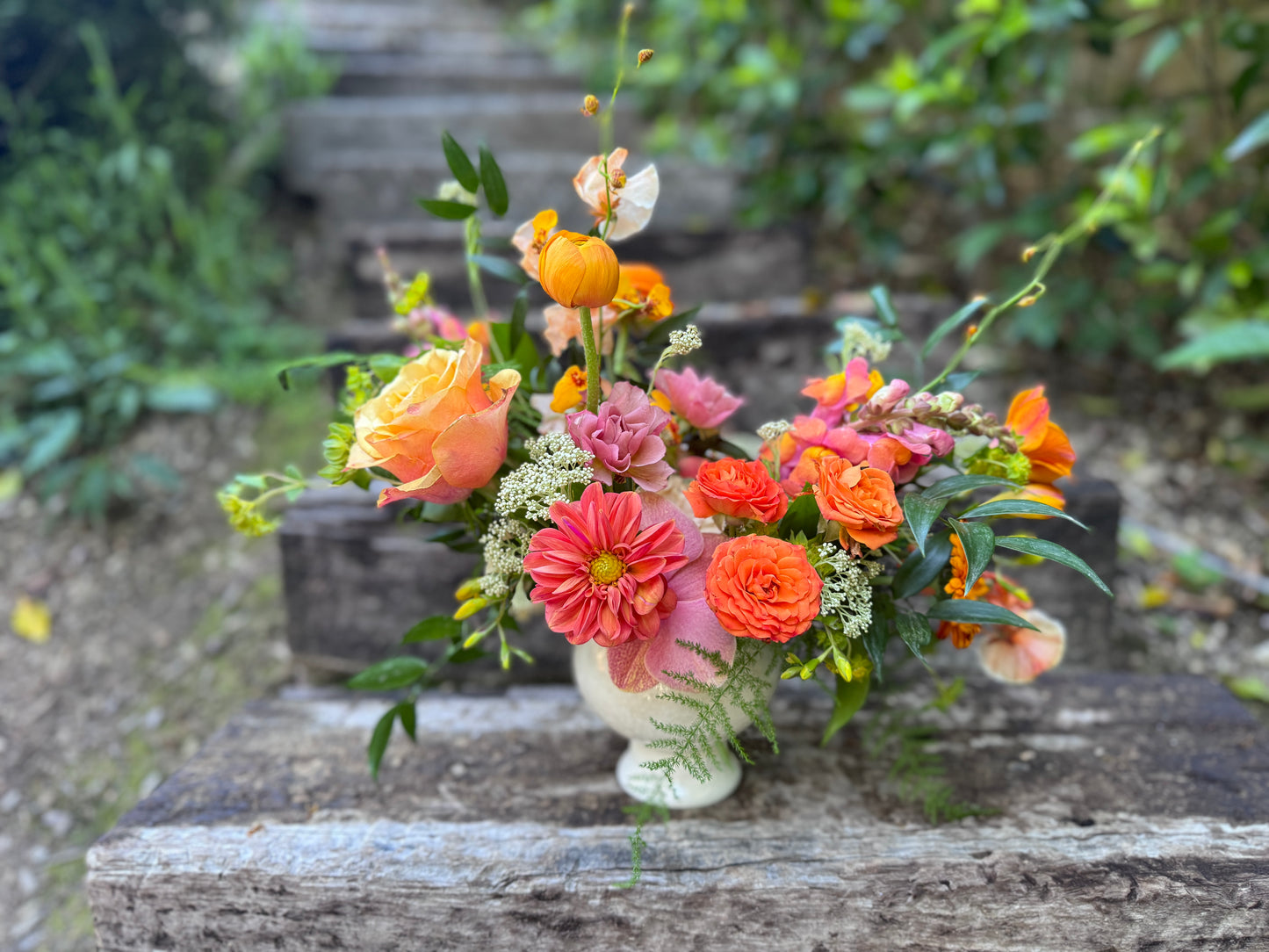 A vibrant floral arrangement featuring orange, pink, and peach flowers with green foliage, presented in a small white ceramic pot on a weathered wooden surface with a blurred garden background.