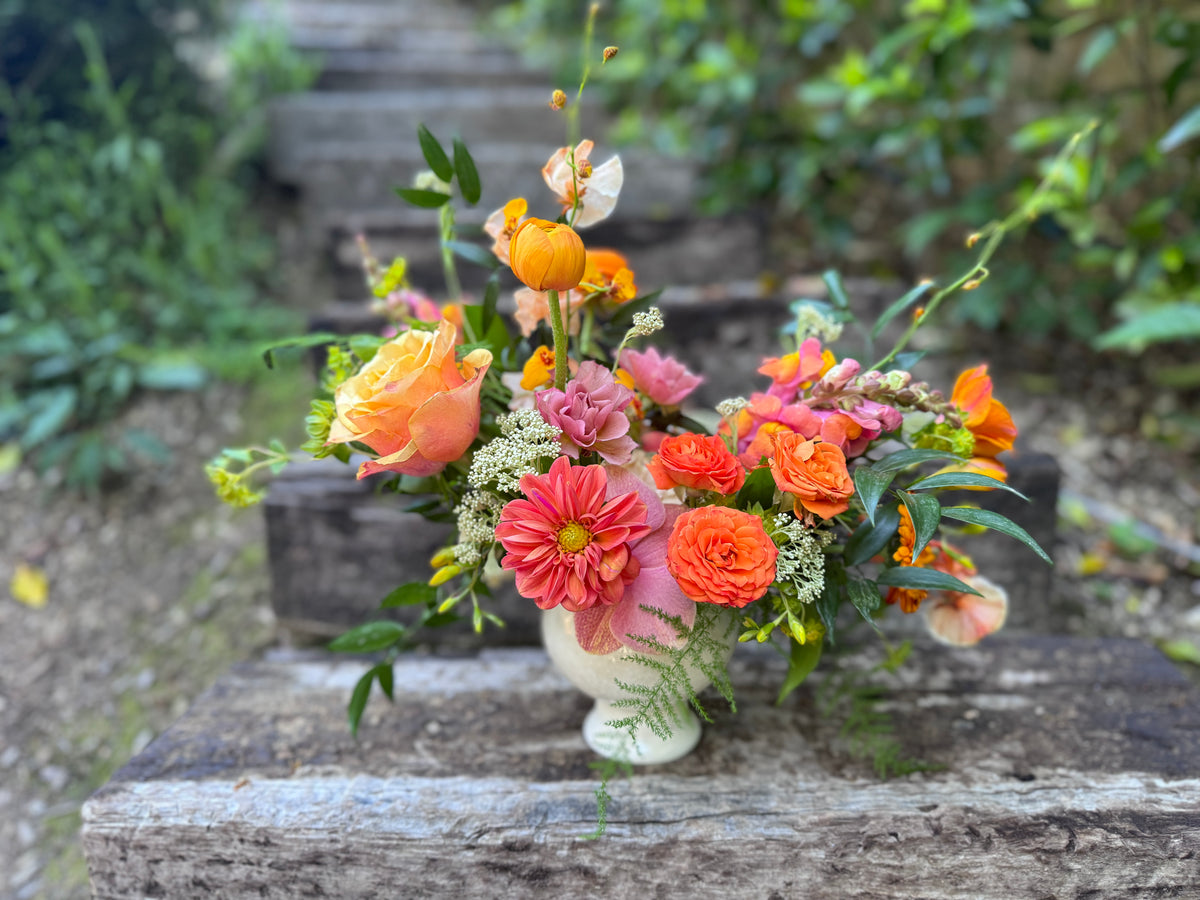 A vibrant floral arrangement featuring orange, pink, and peach flowers with green foliage, presented in a small white ceramic pot on a weathered wooden surface with a blurred garden background.