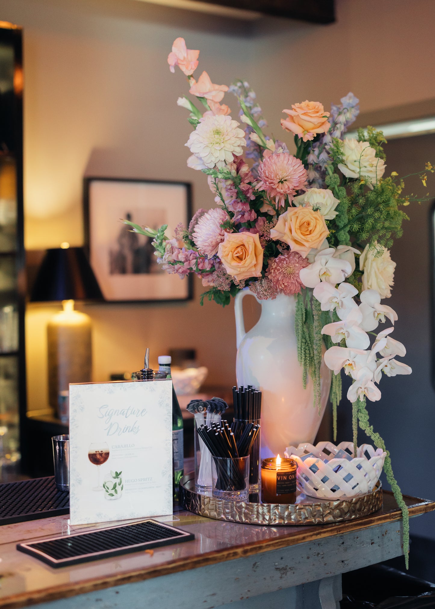 A large floral arrangement in a tall white vase, styled on a bar with candles and a drink menu card in a warmly lit indoor setting.