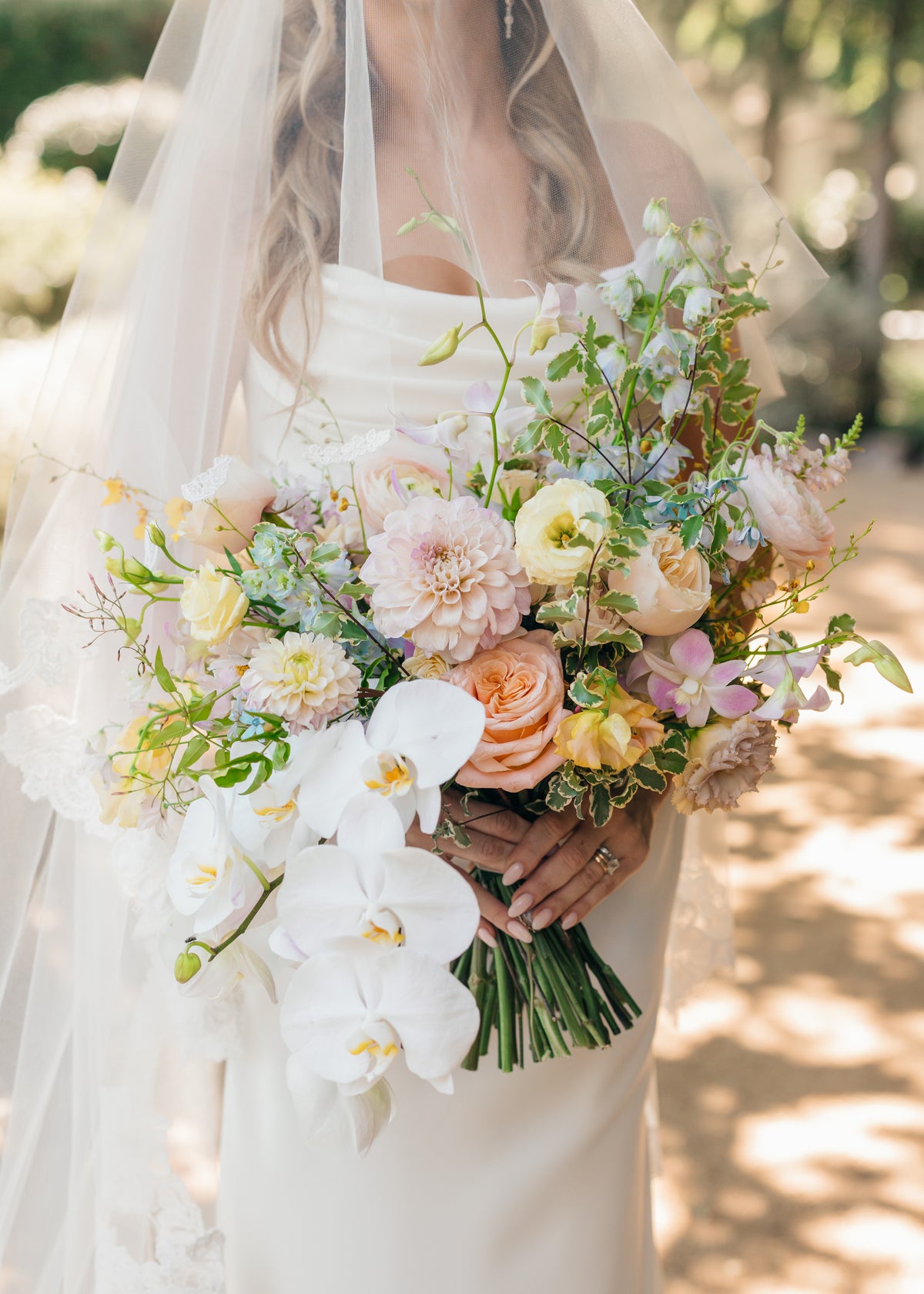 A bride holds a large, lush bridal bouquet featuring cascading white orchids, and premium flowers in pastel shades of cream, butter yellow, peach, blush lavender and light blue at a wedding at The Lodge at Malibou Lake in Agoura Hills, CA 91301.