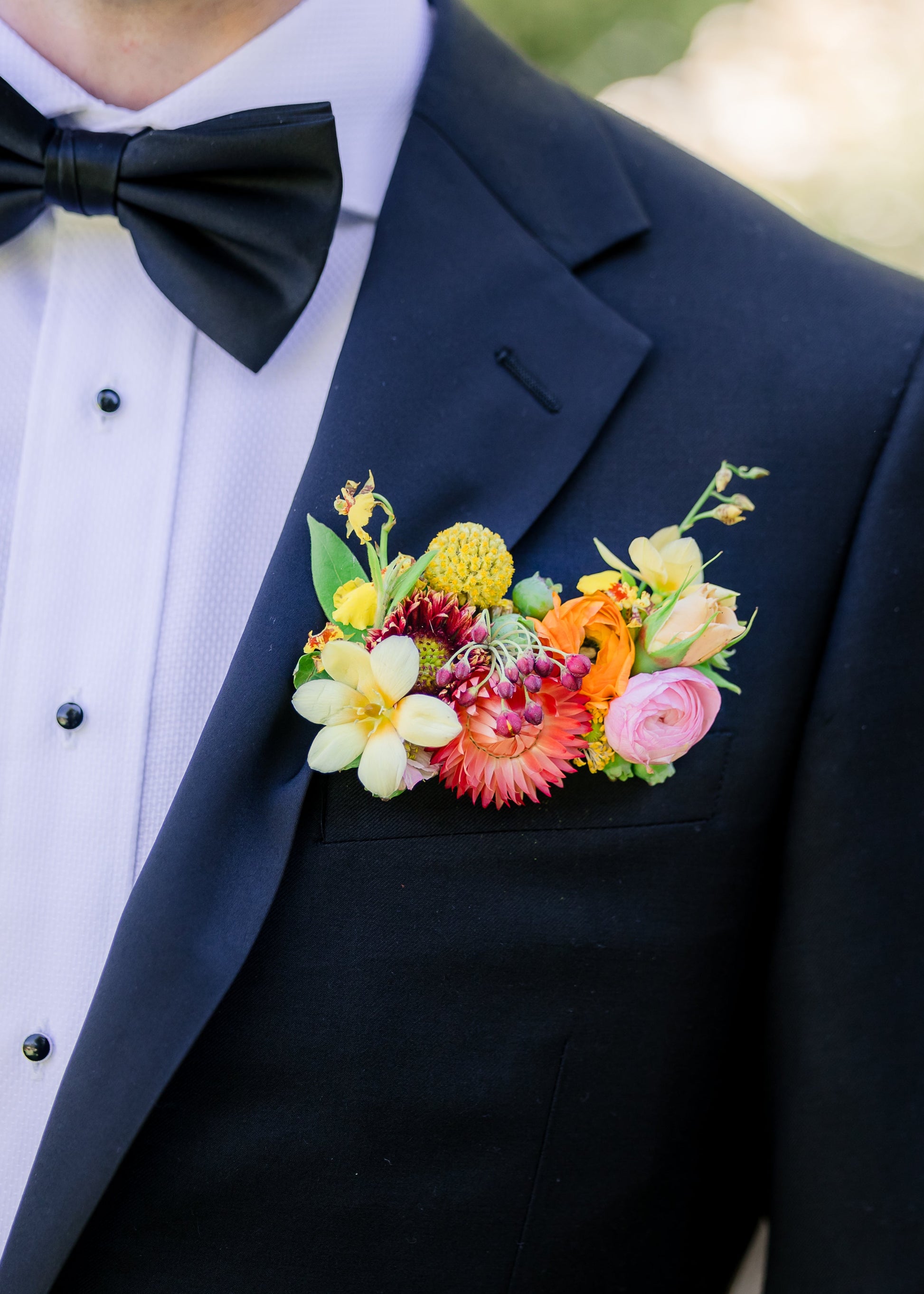 Person wearing a black tuxedo with a colorful floral pocket square boutonniere and bow tie.