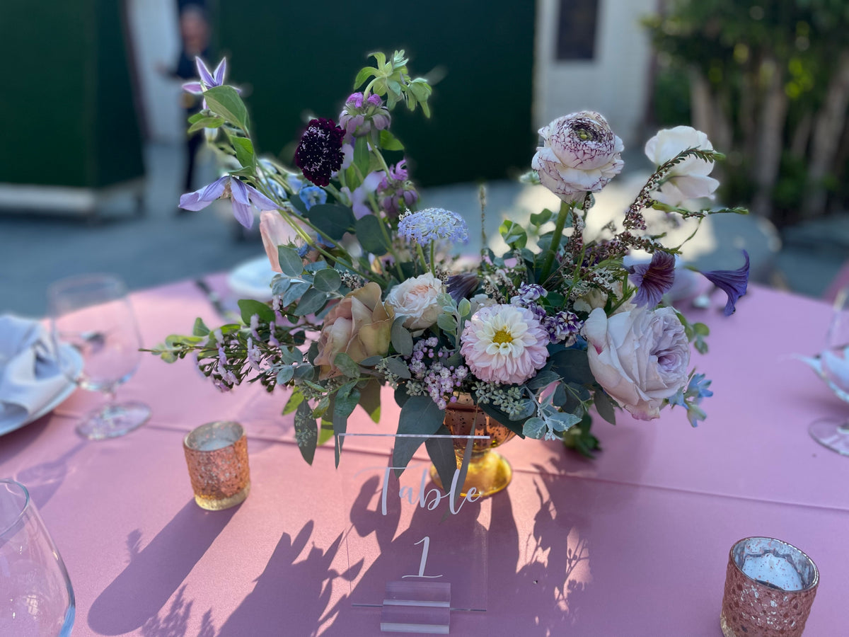 A floral table centerpiece with white, lavender, and green flowers arranged on a pink tablecloth, with table number 1 and small gold candle holders visible.