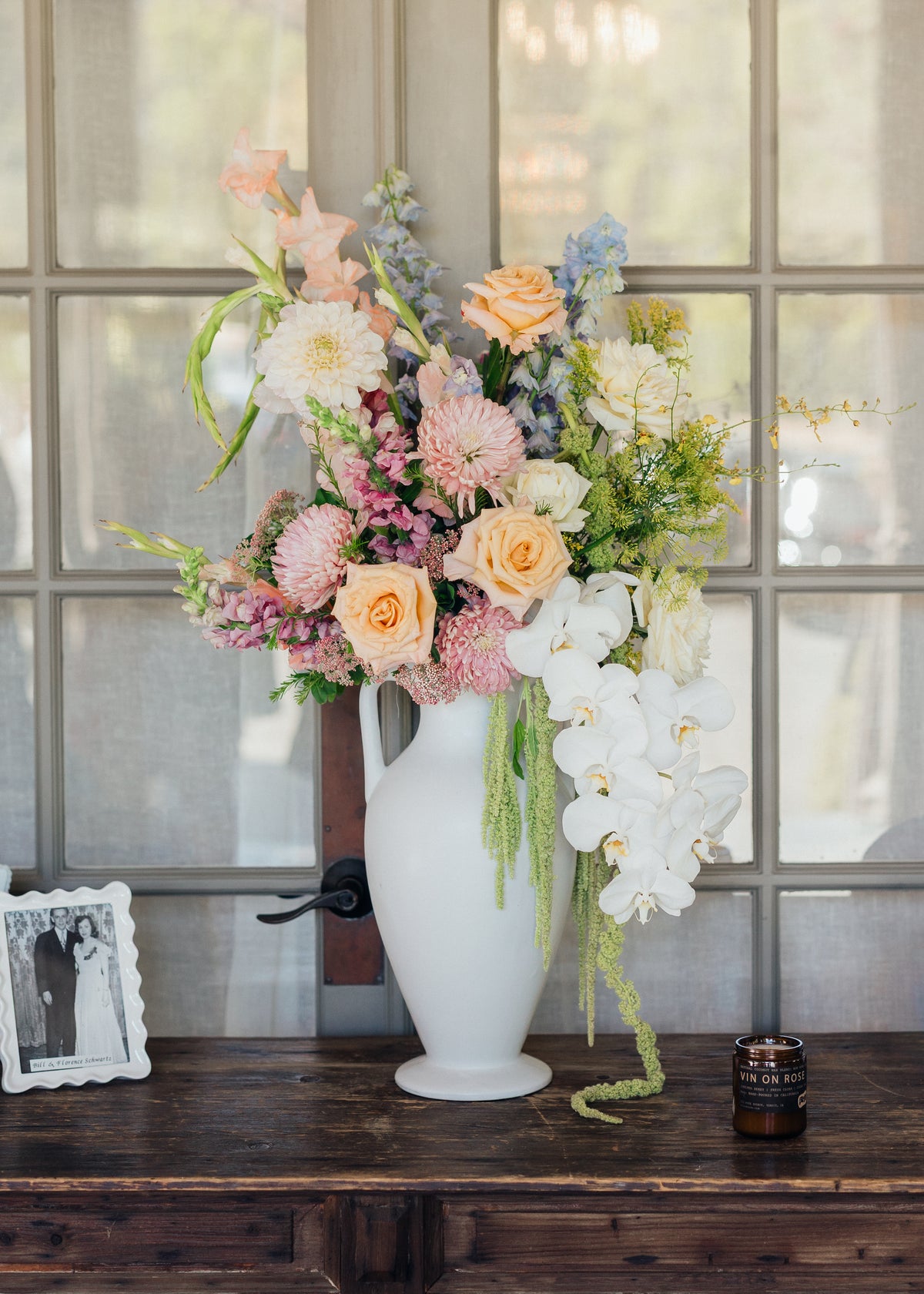 A large white ceramic vase filled with a lush pastel floral arrangement, featuring white phalaenopsis orchids and hanging green amaranthus.