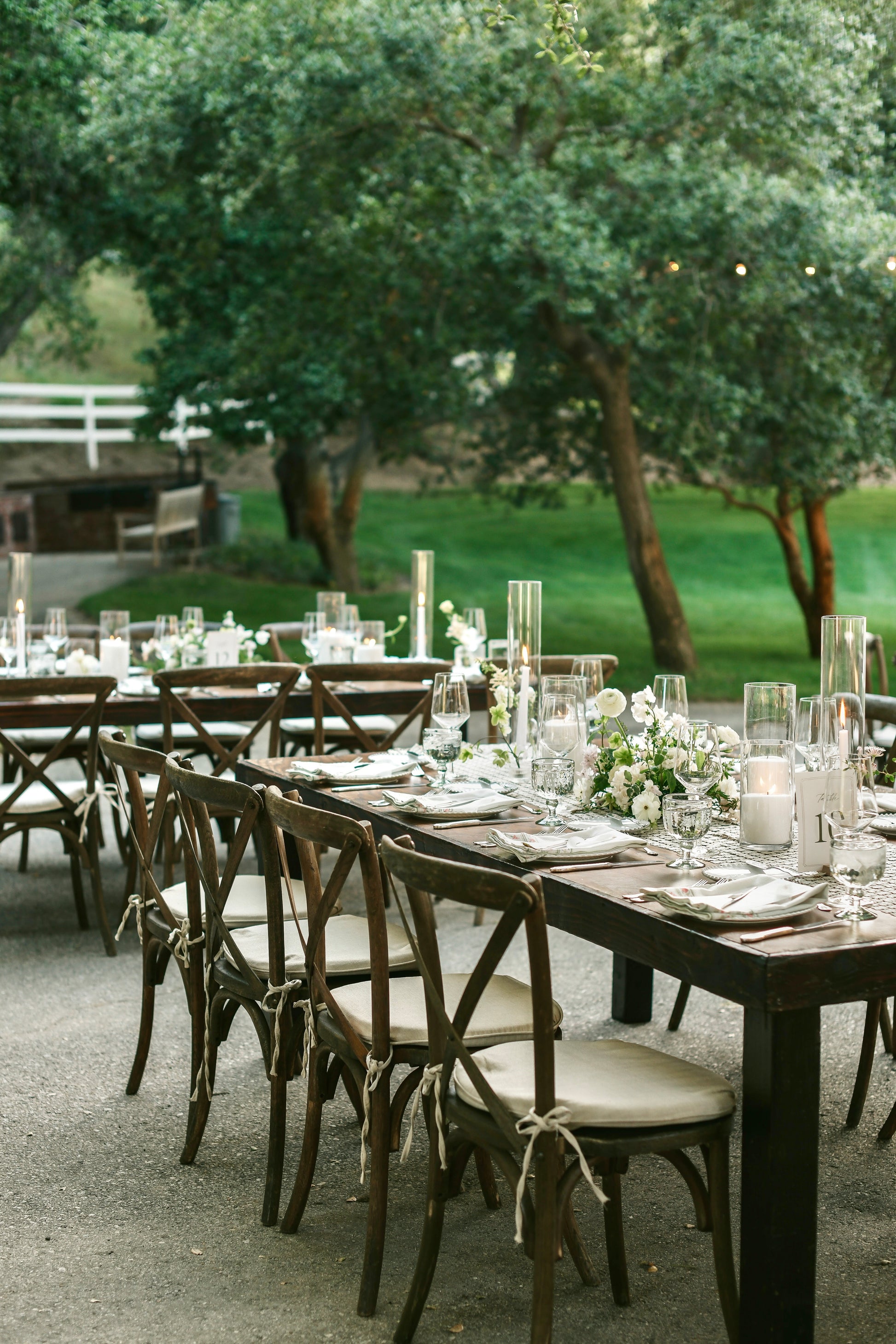 Outdoor wedding reception tables set with dark wood dining chairs, white floral centerpieces, and clear glass candlesticks and water glasses, with green trees and grass in the background.