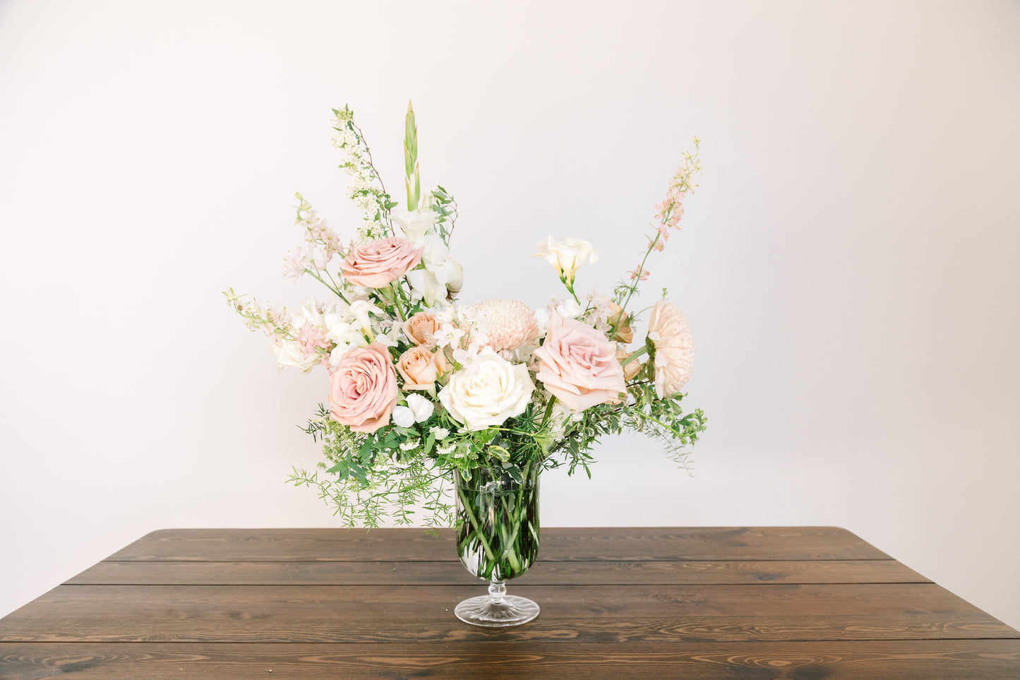 A floral arrangement of pink and white roses with green foliage in a clear glass vase, set on a dark wooden table against a plain light background.