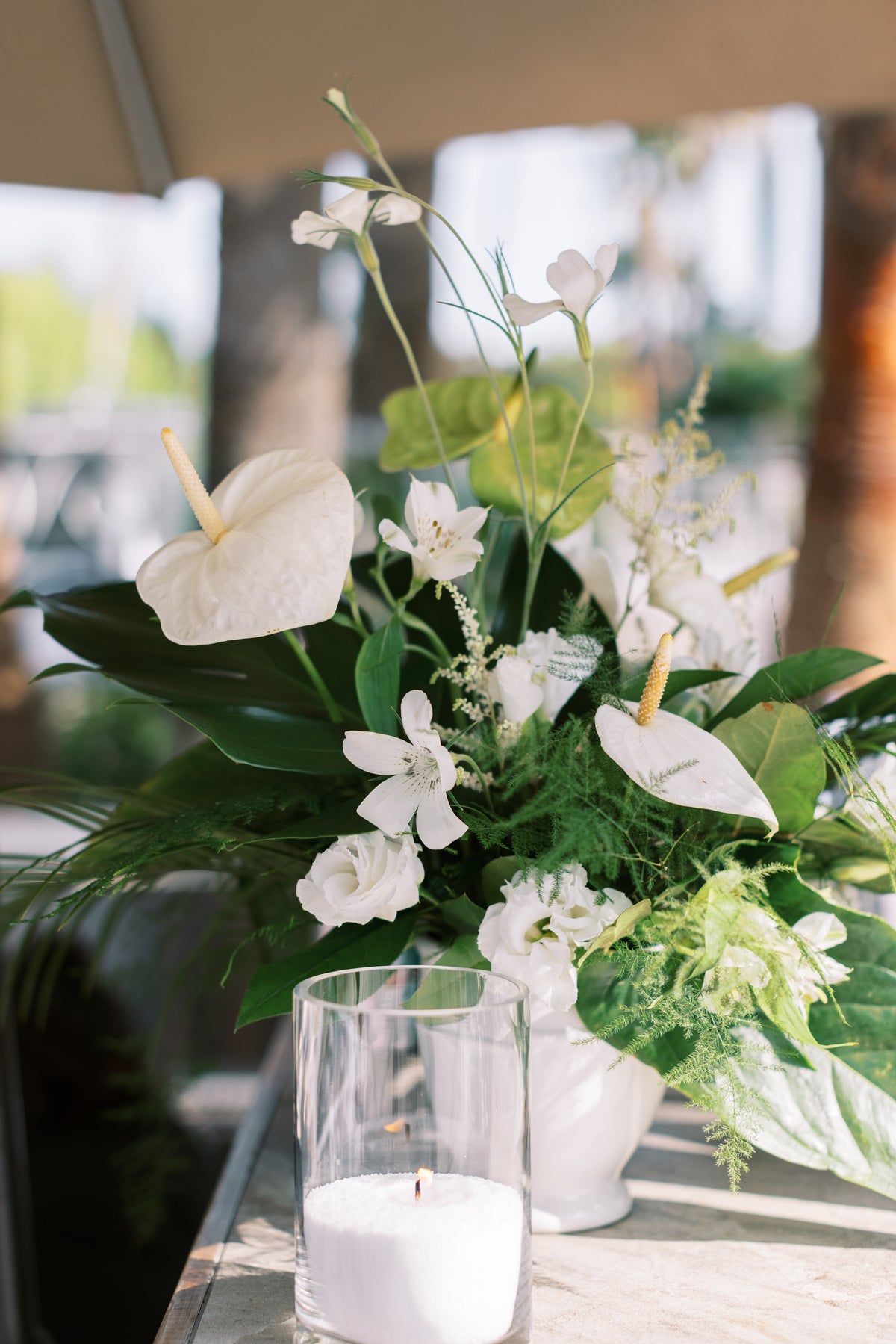 A floral centerpiece arrangement with white flowers and green foliage in a clear glass vase on a table, with a white candle in front and a softly blurred outdoor background.