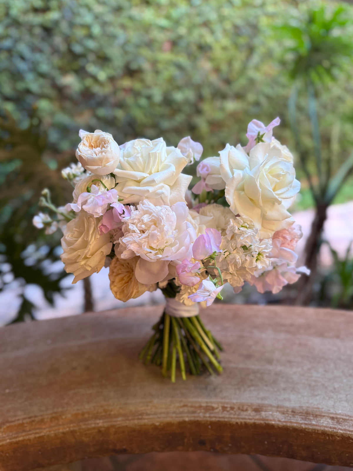 A bouquet of cream, blush pink, and light pink flowers, including roses, sweet peas and peonies, tied with a white ribbon.