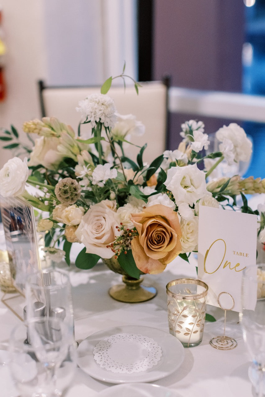 A wedding table centerpiece featuring a lush floral arrangement with white and golden mustard roses, green foliage, and a gold compote vase, styled with clear glassware, a lit votive candle, and a white place card reading 'Table One'.